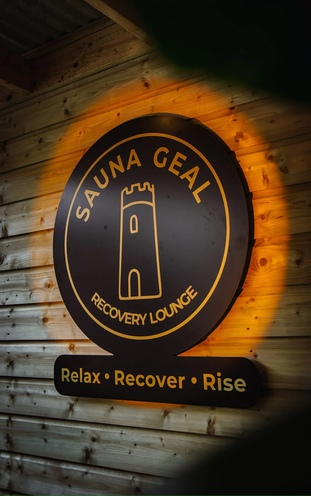 The round Sauna Geal sign lit from behind on a timber wall, reading Relax, Recover, Rise